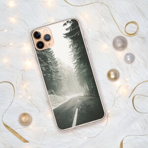 Op de afbeelding: Een transparante telefoonhoes met een winters boslandschap. De afbeelding toont een weg die door een besneeuwd bos slingert met hoge bomen aan beide kanten. De hoes heeft een zwarte camera lens uitsparing.