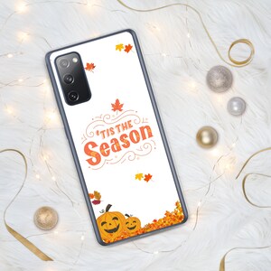 May include: A white phone case with a fall theme. The case features the text "'Tis the Season" in orange, along with illustrations of pumpkins and autumn leaves. The case is surrounded by a white fluffy surface and decorative lights.