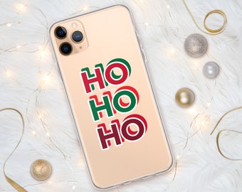 May include: Transparent phone case featuring the words "HO HO HO" in red and green, a festive design for Christmas. The phone case is on a white fluffy surface with decorative baubles and fairy lights.