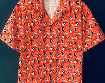 Peut inclure: Chemise orange à manches courtes avec un motif répété de la lettre "I" et du mot "ILLINOIS" en blanc et bleu marine. La chemise a un col et est présentée sur un cintre noir.