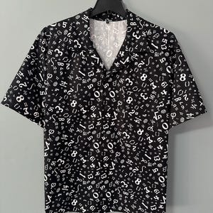 May include: Black button-down shirt with a white number and symbol pattern. The short-sleeved shirt is hanging on a black hanger against a gray background. The shirt has a V-neck collar.