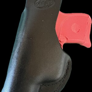 May include: A black leather holster with a red handgun partially inserted. The holster has a smooth, curved design with visible stitching. The handgun's grip is textured.