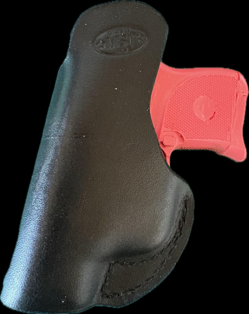 May include: A black leather holster with a red handgun partially inserted. The holster has a smooth, curved design with visible stitching. The handgun's grip is textured.