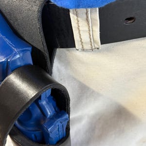 Op de afbeelding: Een zwarte leren holster met een blauw object erin. De holster is bevestigd aan een zwarte riem met een gesp. Blauwe stof is zichtbaar boven de holster.