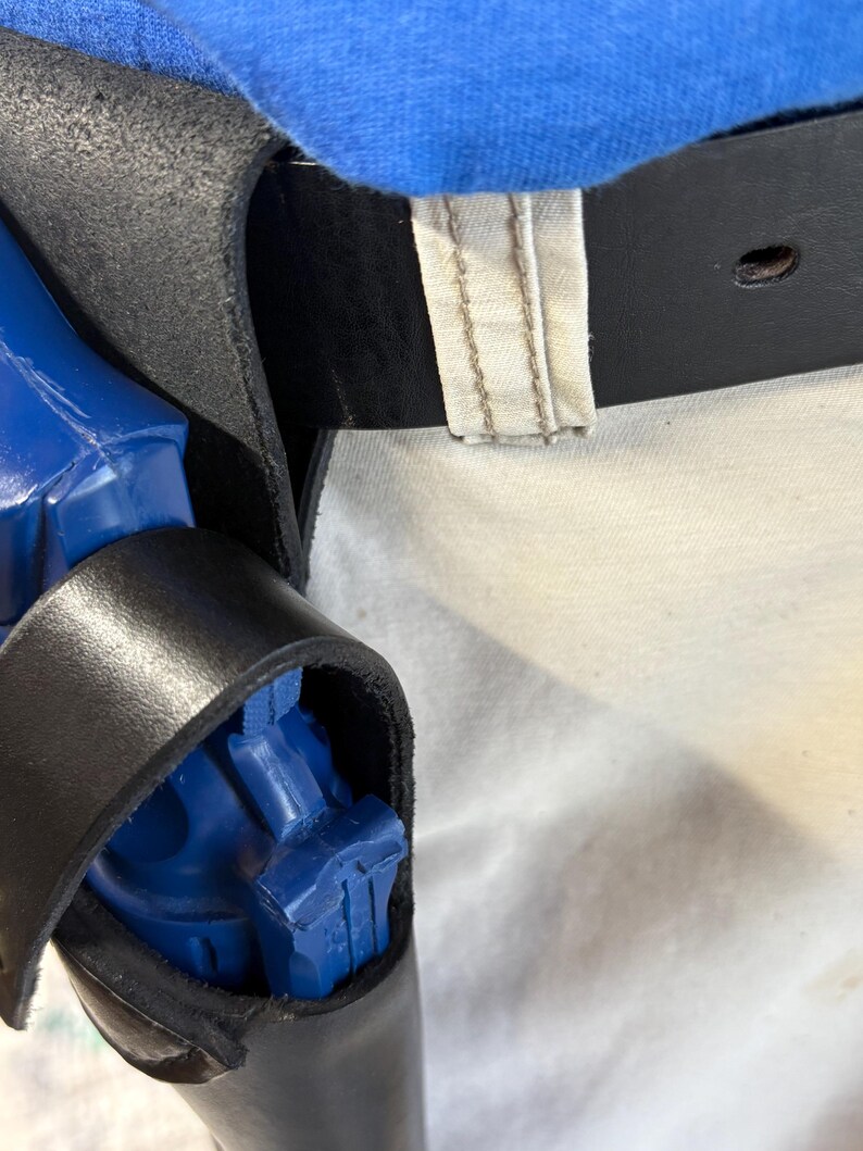 Op de afbeelding: Een zwarte leren holster met een blauw object erin. De holster is bevestigd aan een zwarte riem met een gesp. Blauwe stof is zichtbaar boven de holster.