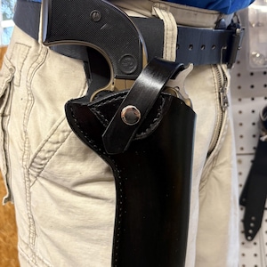 K&ouml;nnte beinhalten: Eine schwarze Lederholster mit einem silbernen Druckknopf, die eine Schusswaffe h&auml;lt. Die Holster ist an einem schwarzen G&uuml;rtel befestigt, der mit khakifarbener Hose getragen wird. Die Schusswaffe hat einen schwarzen Griff und einen silbernen Lauf.