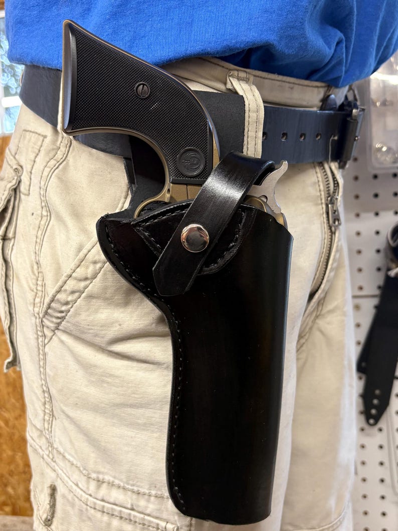 K&ouml;nnte beinhalten: Eine schwarze Lederholster mit einem silbernen Druckknopf, die eine Schusswaffe h&auml;lt. Die Holster ist an einem schwarzen G&uuml;rtel befestigt, der mit khakifarbener Hose getragen wird. Die Schusswaffe hat einen schwarzen Griff und einen silbernen Lauf.