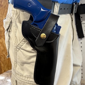 Op de afbeelding: Een zwarte leren holster met een goudkleurige knop die een blauw pistool vastzet. De holster is bevestigd aan een zwarte riem die wordt gedragen met een kaki broek. Het pistool is gedeeltelijk zichtbaar in de holster.