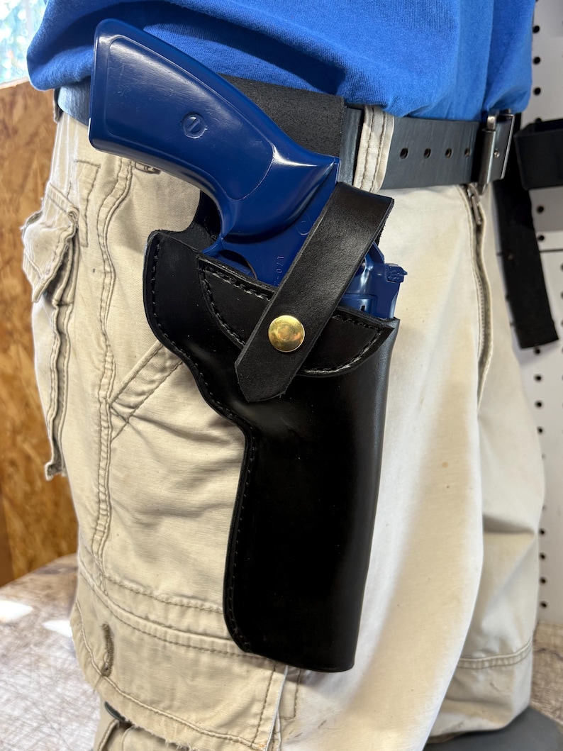 Op de afbeelding: Een zwarte leren holster met een goudkleurige knop die een blauw pistool vastzet. De holster is bevestigd aan een zwarte riem die wordt gedragen met een kaki broek. Het pistool is gedeeltelijk zichtbaar in de holster.