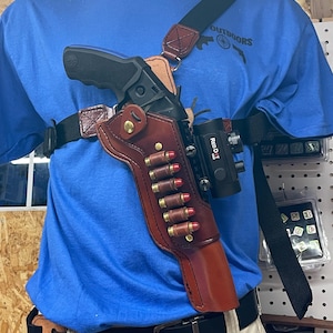 May include: A brown leather holster with a red dot sight attached to a revolver. The holster is worn on a person's shoulder with a black strap. The revolver has a black handle and a silver barrel. The holster has a magazine with six bullets.