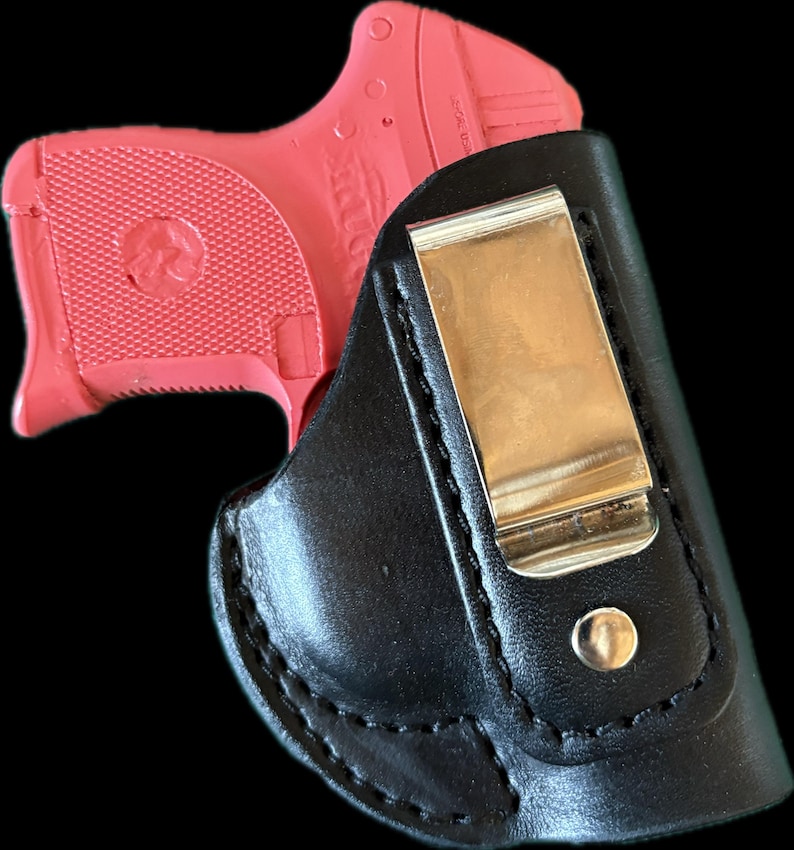 May include: A bright pink handgun partially visible inside a black leather holster. The holster has a gold-colored metal clip and a snap closure. The handgun's grip has a textured pattern.