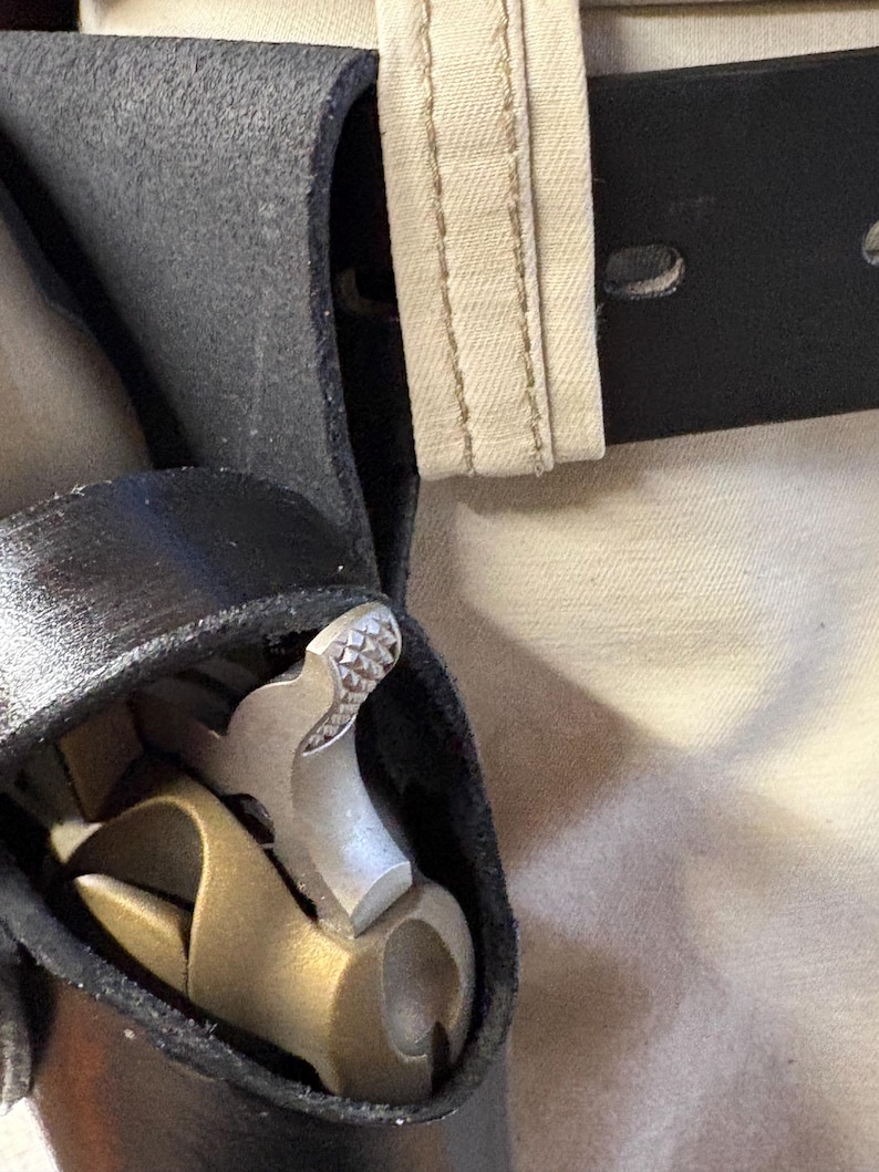 K&ouml;nnte beinhalten: Nahaufnahme eines schwarzen Leder-Werkzeugg&uuml;rtels mit einem silbernen und Messinghammer und anderen Werkzeugen. Der G&uuml;rtel wird mit einem schwarzen G&uuml;rtel und einer beigefarbenen Hose getragen.