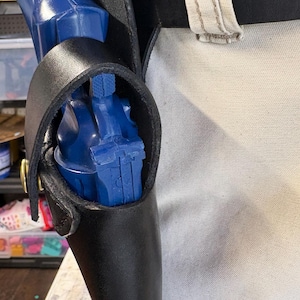 Op de afbeelding: Een zwart leren holster met een blauw pistool erin. Het holster heeft een lus voor een riem en is vastgezet met een messingkleurige klinknagel. De achtergrond is een lichtgekleurde stof.