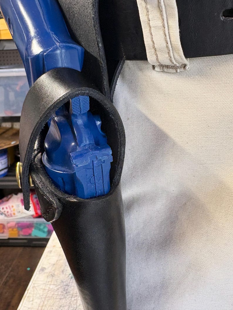 Op de afbeelding: Een zwart leren holster met een blauw pistool erin. Het holster heeft een lus voor een riem en is vastgezet met een messingkleurige klinknagel. De achtergrond is een lichtgekleurde stof.