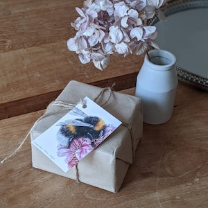 Puede incluir: Una caja de regalo envuelta en papel marrón con una etiqueta que muestra una ilustración pintada a mano de un abejorro sobre flores rosas. La etiqueta dice "Handmade with Care".