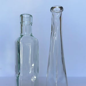 May include: Two clear glass bottles, one with a square shape and the other with a triangular shape. Both bottles are empty and have a smooth, polished surface.