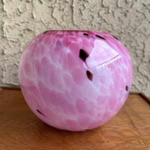 Puede incluir: Un jarr&oacute;n de vidrio rosa redondo con un patr&oacute;n moteado de manchas blancas y marr&oacute;n oscuro. El jarr&oacute;n tiene un acabado liso y brillante y se asienta sobre una superficie de madera. El jarr&oacute;n es un art&iacute;culo decorativo.