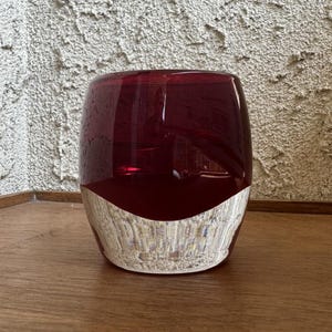 May include: A cylindrical glass vase with a deep red upper half and a textured, light-colored lower half. The vase sits on a wooden surface, and the background is a textured wall.