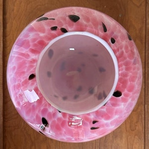 Puede incluir: Un cuenco de vidrio rosa con un patr&oacute;n moteado de puntos negros. El cuenco tiene una forma ancha y redondeada con un borde blanco y una abertura circular m&aacute;s peque&ntilde;a en el centro. El cuenco est&aacute; sobre una superficie de madera.