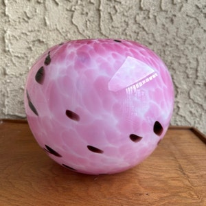 Puede incluir: Un jarr&oacute;n de vidrio soplado a mano, de forma redonda, con un dise&ntilde;o moteado en rosa y blanco. Manchas de color marr&oacute;n oscuro est&aacute;n dispersas por la superficie. El jarr&oacute;n est&aacute; sobre una superficie de madera.