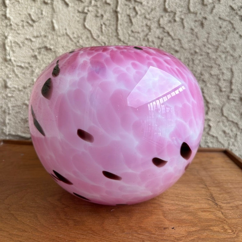 Puede incluir: Un jarr&oacute;n de vidrio soplado a mano, de forma redonda, con un dise&ntilde;o moteado en rosa y blanco. Manchas de color marr&oacute;n oscuro est&aacute;n dispersas por la superficie. El jarr&oacute;n est&aacute; sobre una superficie de madera.