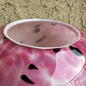 Puede incluir: Un jarr&oacute;n de cristal rosa con borde blanco y detalles morados oscuros. El jarr&oacute;n tiene un patr&oacute;n moteado y una abertura ancha. El interior del jarr&oacute;n es de un tono rosa m&aacute;s claro.