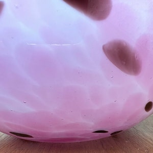 Puede incluir: Un cuenco redondo de vidrio rosa claro con un dise&ntilde;o moteado. El cuenco tiene una abertura ancha y est&aacute; decorado con manchas marrones oscuras. El vidrio tiene un aspecto texturizado y est&aacute; sobre una superficie de madera.