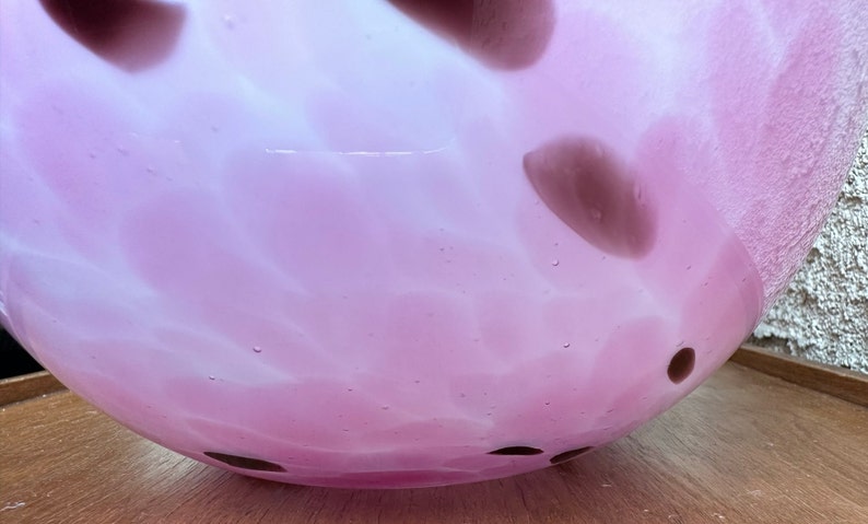 Puede incluir: Un cuenco redondo de vidrio rosa claro con un dise&ntilde;o moteado. El cuenco tiene una abertura ancha y est&aacute; decorado con manchas marrones oscuras. El vidrio tiene un aspecto texturizado y est&aacute; sobre una superficie de madera.