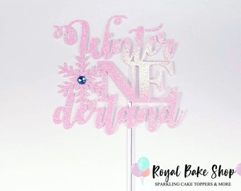 Könnte beinhalten: Rosa glitzernder Tortendekor mit den Worten "Winter One" und einer Schneeflocke mit einem blauen Stein in der Mitte. Der Dekor ist auf einem weißen Hintergrund.