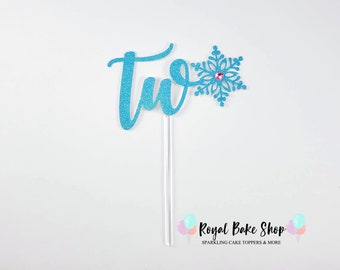 Könnte beinhalten: Blauer Glitzer-Kuchentopper mit dem Wort "zwei" und einem Schneeflocken-Design. Die Schneeflocke hat einen rosafarbenen Strassstein in der Mitte. Der Topper ist auf einem weißen Stiel. Der Text "Royal Bake Shop Sparkling Cake Toppers & More" befindet sich am unteren Rand des Bildes.