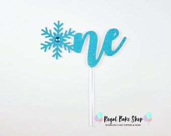 Könnte beinhalten: Blauer Glitzer-Kuchentopper mit einer Schneeflocke und dem Wort "eins" in Kursivschrift. Royal Bake Shop Sparkling Cake Toppers & More.