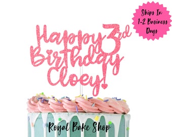 Könnte beinhalten: Rosa glitzernder Tortendekor mit der Aufschrift "Happy 3rd Birthday Cloey!" Der Dekor befindet sich auf einem hellblauen und rosa glasierten Kuchen mit Streuseln.