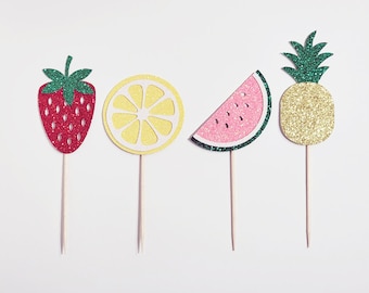 Könnte beinhalten: Vier glitzernde Obst-Kuchentopper: eine rote Erdbeere, eine gelbe Zitrone, ein rosa Wassermelonenstück und eine goldene Ananas. Jeder Topper ist auf einem Holzstiel.
