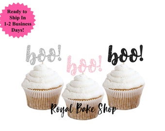Könnte beinhalten: Drei Cupcakes mit weißem Zuckerguss, jeweils gekrönt von einem glitzernden "boo!" Tortenaufsatz in Silber, Rosa und Schwarz. Die Cupcakes befinden sich in braunen Papierförmchen. Der Text "Royal Bake Shop" steht unten.