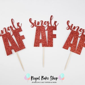 May include: Three red glitter cake toppers with the text "Single AF" on each. The toppers are on wooden sticks.