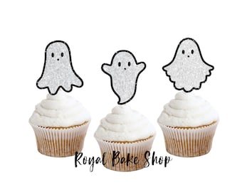 Könnte beinhalten: Drei Cupcakes mit weißem Zuckerguss und Geisterdekorationen. Die Geister-Topper sind silberfarben mit schwarzen Umrissen. Der Text "Royal Bake Shop" befindet sich am unteren Rand des Bildes.