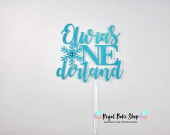 Könnte beinhalten: Blauer Glitzer-Kuchentopper mit dem Text "Olivia's One derland" und einer Schneeflocke mit einem blauen Stein in der Mitte.