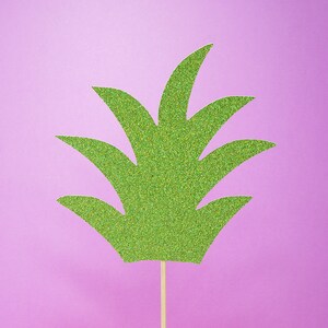 Puede incluir: Un recorte de pi&ntilde;a verde con purpurina sobre un fondo morado claro. La pi&ntilde;a est&aacute; en un palo de madera.