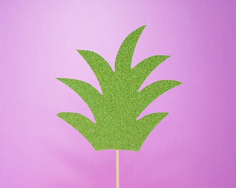 Puede incluir: Un recorte de pi&ntilde;a verde con purpurina sobre un fondo morado claro. La pi&ntilde;a est&aacute; en un palo de madera.