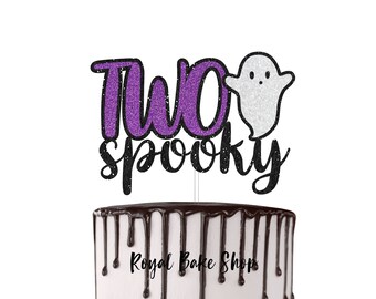 Könnte beinhalten: Ein Tortenaufsatz mit den Worten "Two Spooky" in lila und schwarzem Glitzer. Ein weißer Glitzergeist befindet sich neben den Worten. Der Kuchen hat eine weiße Glasur mit Schokoladentropfen.
