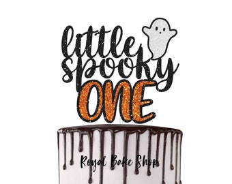 Könnte beinhalten: Ein Halloween-Kuchenaufsatz mit den Worten "little spooky ONE" in schwarzem und orangefarbenem Glitzer. Ein weißer Glitzergeist befindet sich über dem Wort "spooky". Der Kuchen hat eine weiße Glasur mit Schokoladentropfen. Der Text "Royal Bake Shop" steht unten.