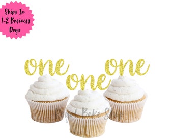 Könnte beinhalten: Drei Cupcakes mit weißem Zuckerguss und goldenen "one"-Toppern. Die Cupcakes befinden sich in braunen Papierförmchen. Das Bild hat einen weißen Hintergrund. Der Text "Royal Bake Shop" befindet sich in der unteren rechten Ecke.