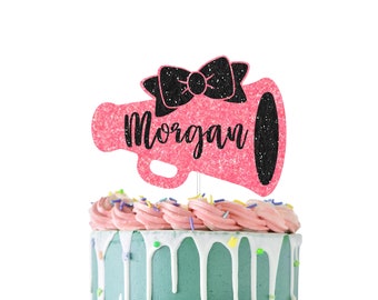 Könnte beinhalten: Ein rosa glitzernder Megaphon-Kuchentopper mit einer schwarzen Schleife und dem Namen "Morgan" darauf.