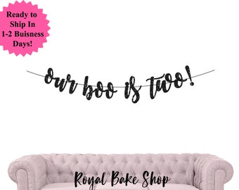 Puede incluir: Pancarta de purpurina negra y blanca que dice "our boo is two!" colgada sobre un sofá rosa claro con el texto "Royal Bake Shop" en la parte delantera.