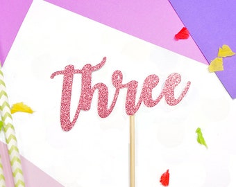 May include: Pink glitter cake topper with the word "Three" in a cursive font. The topper is on a white background with colorful confetti.