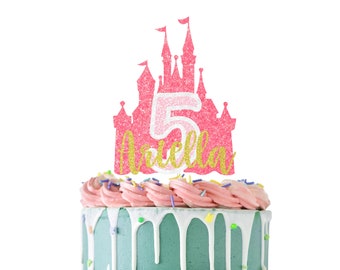 Könnte beinhalten: Ein rosa glitzernder Schloss-Kuchenaufsatz mit der Zahl 5 und dem Namen "Ariella" in goldglitzer. Der Kuchen hat rosa Zuckerguss, Streusel und eine hellblaue Basis mit weißen Zuckerguss-Tropfen. Ein festlicher Geburtstagskuchen.