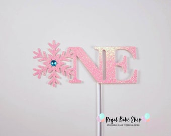Könnte beinhalten: Ein rosa glitzernder Tortendekor mit der Zahl eins und einer Schneeflocke. Die Schneeflocke hat einen blauen Strassstein in der Mitte. Der Dekor ist auf einem weißen Hintergrund. Royal Bake Shop Sparkling Cake Toppers & More.