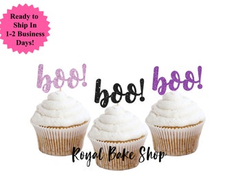Könnte beinhalten: Drei Cupcakes mit weißem Zuckerguss und glitzernden "boo!"-Toppern in Rosa, Schwarz und Lila. Die Cupcakes befinden sich in braunen Papierförmchen. Der Text "Royal Bake Shop" steht unten.