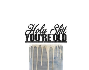 Könnte beinhalten: Ein schwarzer Glitzer-Kuchenaufsatz mit dem Schriftzug "Holy Shit YOU'RE OLD" in einer kräftigen Schriftart. Der Aufsatz befindet sich auf einem Kuchen mit blauer Glasur und weißen Zuckerguss-Tropfen.
