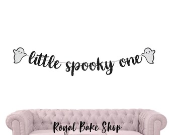 Könnte beinhalten: Ein Banner mit dem Text "little spooky one" in schwarzem Glitzer, flankiert von zwei silbernen Glitzer-Geistern. Das Banner hängt vor einem weißen Hintergrund, über einem rosa Sofa.